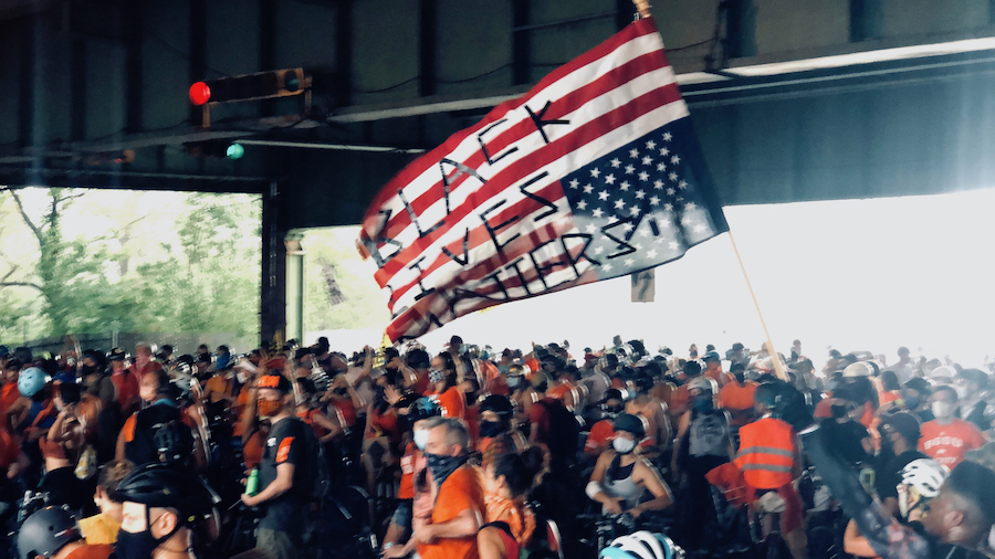 A crowd of protesters stand in the street under an overpass. In the center, someone is holding an upside down American flag that reads "Black Lives Matter."