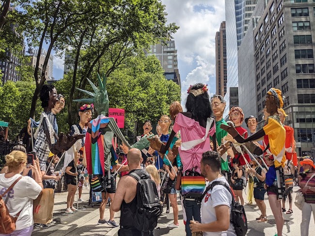 Fotos de um desfile pelas ruas da cidade, muitas pessoas estão vestindo fantasias de marionetes maiores que a vida