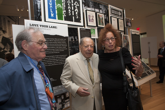 Três indivíduos, dois homens, à esquerda, e uma mulher, à direita, em frente a uma das exposições de estudo de caso na exposição "Activist New York"