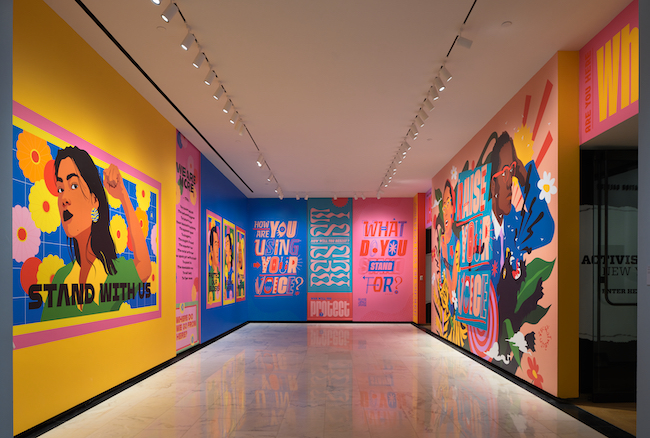 Vue de l'installation "Raise Your Voice", avec des peintures murales aux couleurs vives de portraits et un texte d'accompagnement.