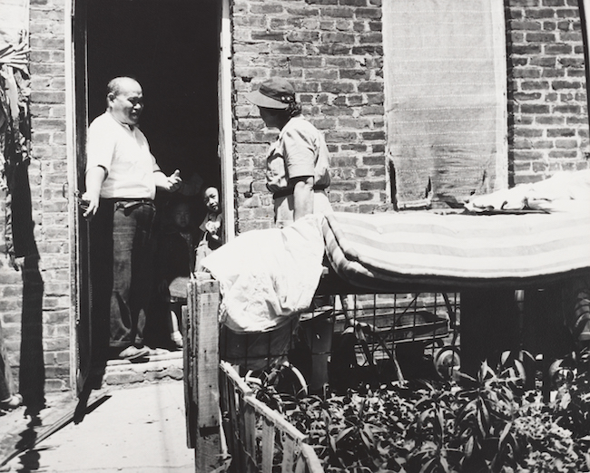 A man stands in a doorway with a small child. A woman is speaking to them from outside of a brick building.