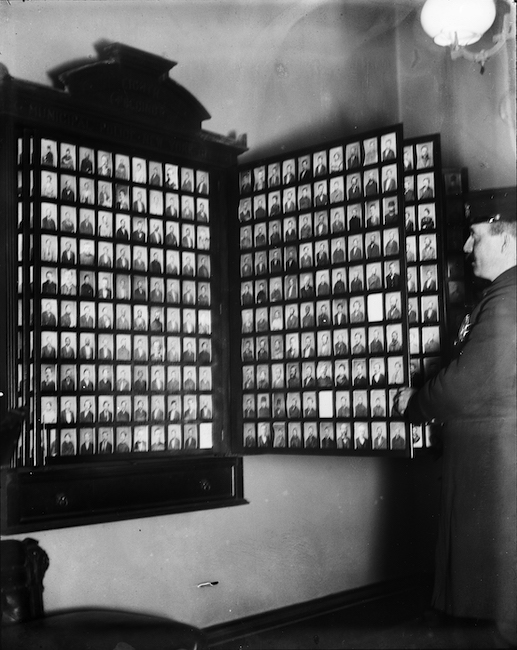 Man looking at a wall case with many tiny photographs of other individuals arranged in a grid.