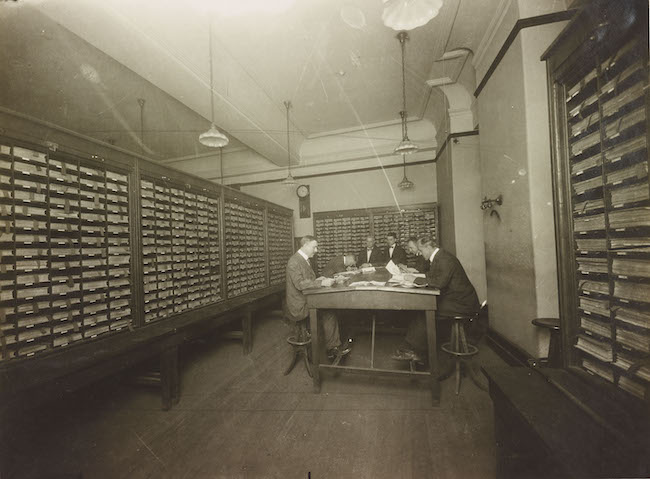 Fotografia mostrando homens olhando para documentos sobre uma mesa, cercados por uma parede forrada de prateleiras de pequenas gavetas.