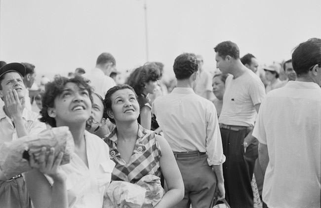 Black & white photograph of a group of people standing outside. Two on the lower left, facing the camera, are staring at the sky.