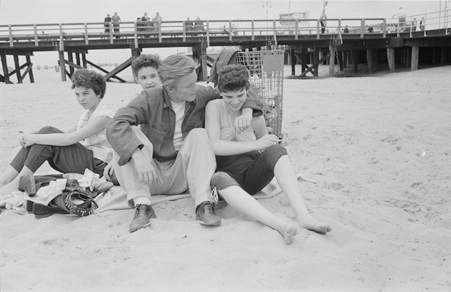 Black & white photograph of a group of four people—three women and one man with his arm around one of the women—sitting on a blanket at the beach. A pier is in the far background.
