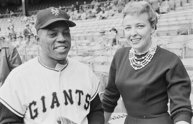 Black & white photograph of an African American man in a Giants baseball uniform and hat stands next to a women in a nice dress and jewelry. A stadium is behind them, out of focus.