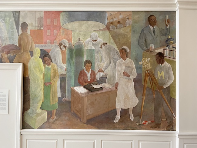 Photograph of a wall mural. In the center is a woman sitting at a desk with a typewriter, a nurse stands in front of it.