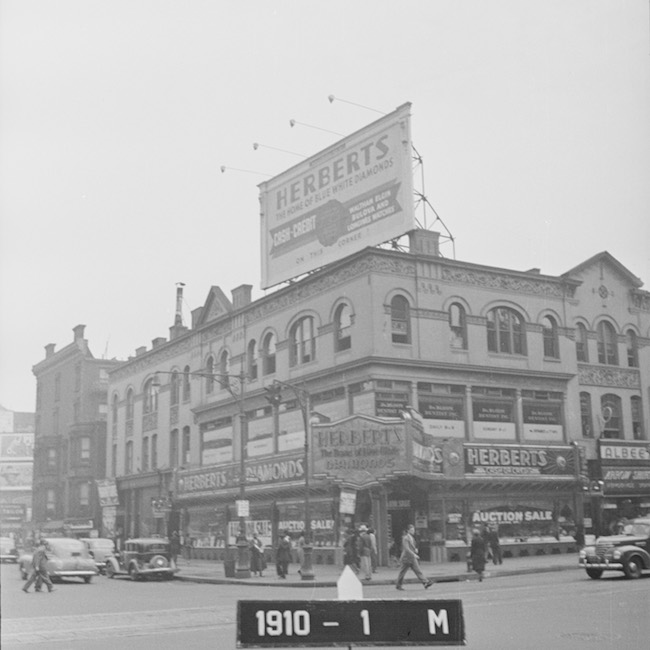 Photographie en noir et blanc d'un coin de rue montrant l'entreprise avec un grand panneau d'affichage sur le dessus pour "Herberts".