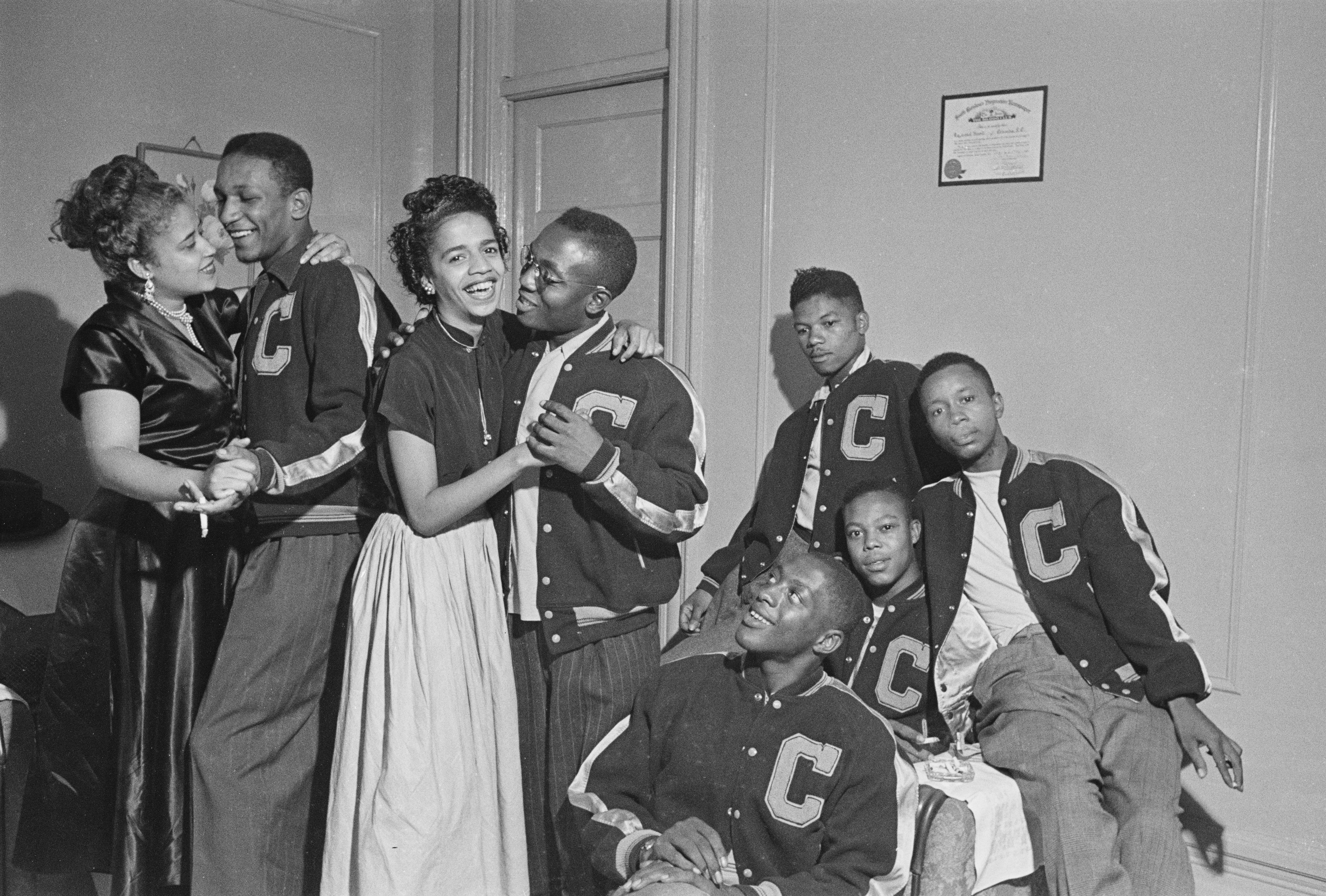 A group of young men and women hang out in a room. Two couples are dancing and smiling. The men are each wearing matching jackets with the letter “C” on the front.