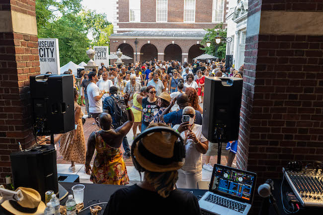 Vista de personas bailando en el patio del Museo en la entrada principal. El fotógrafo estaba parado justo detrás del DJ, por lo que la parte posterior de la cabeza del DJ está al frente. La gente habla, baila y bebe.