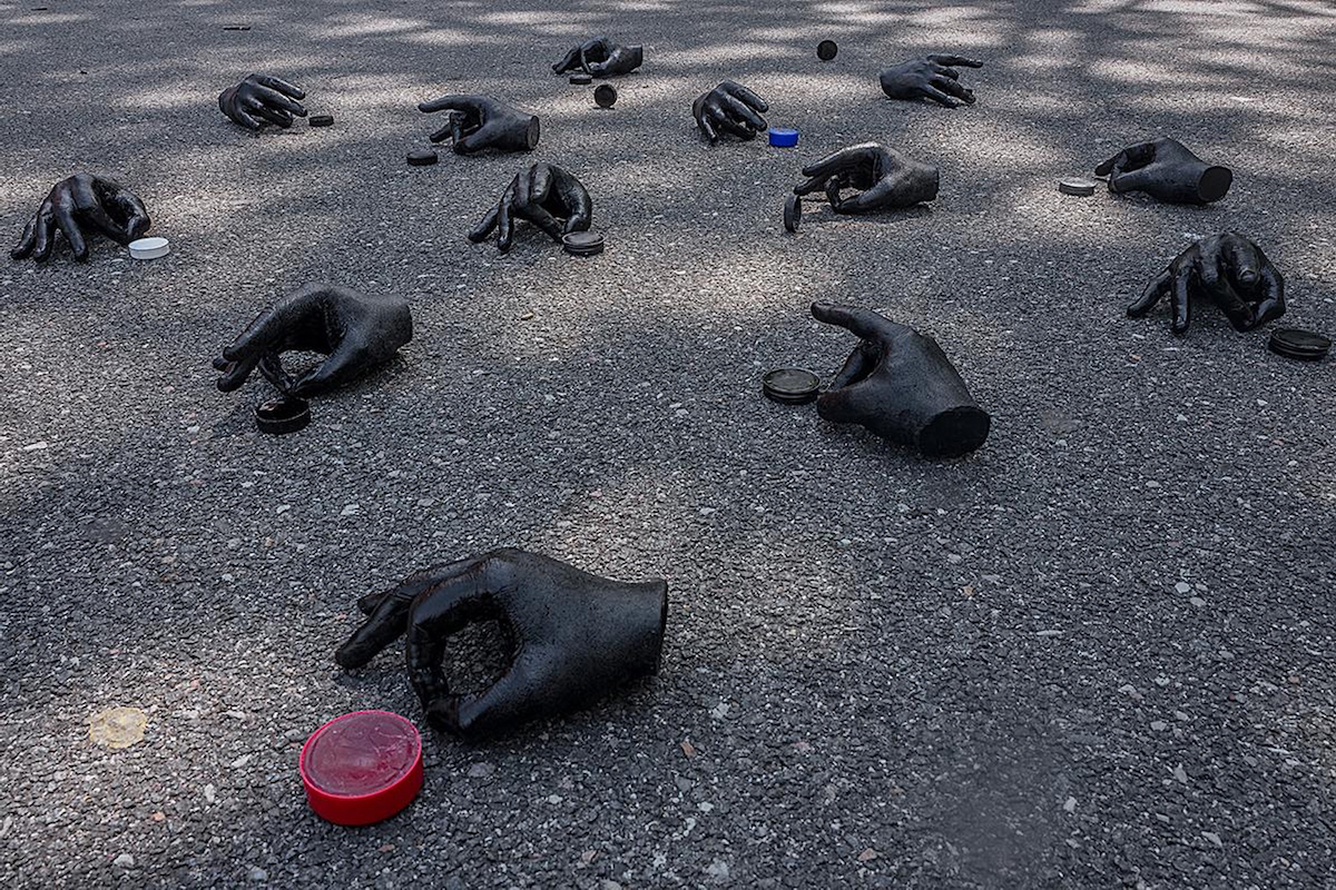 Fotografia de 13 mãos esculpidas, cada uma com vários dedos tocando o polegar. As mãos são colocadas no asfalto, cada uma ao lado de um pequeno círculo de plástico, como uma tampa de garrafa ou disco de várias cores