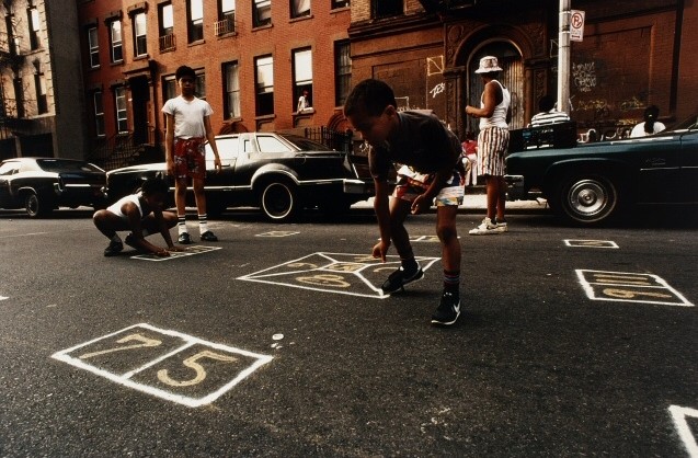 A group of young boys play a game in the middle of a city street - the game involves numbers written in paint or chalk inside of squares or triangles.