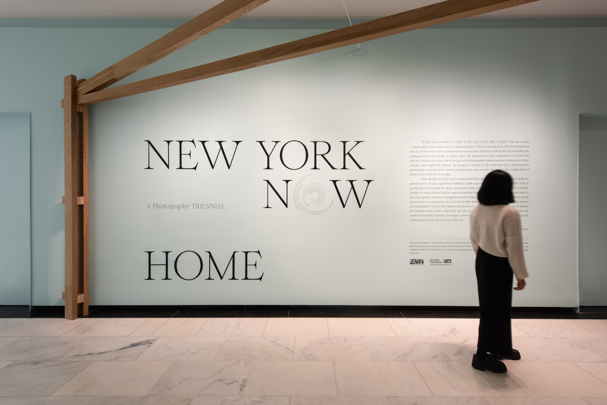 A woman stands in front of the introduction text for the exhibition "New York Now: Home"