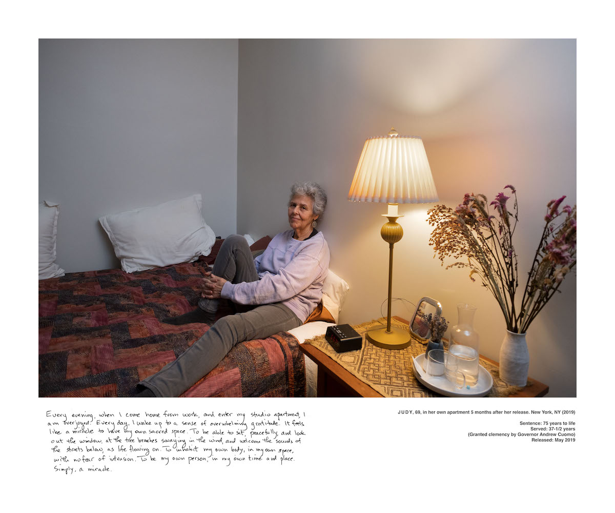 A women sits on a bed with one leg bent up. The light on the bed-side table to her left is on.