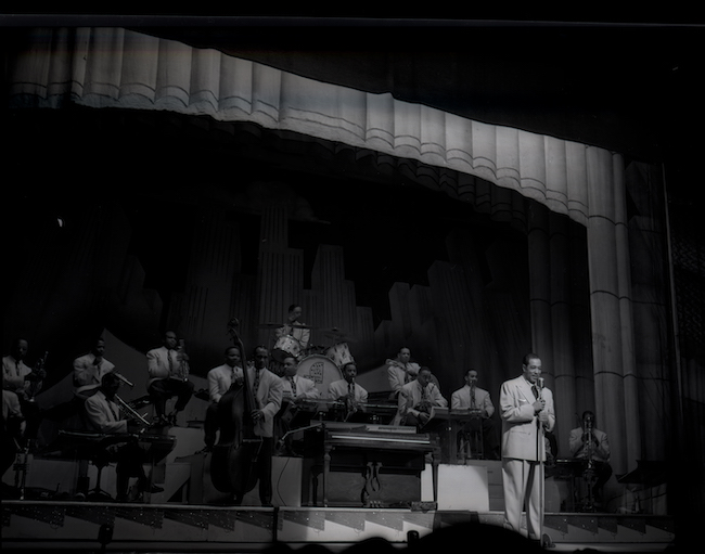 Photograph of a group of musicians on a stage with Duke Ellington in front at the mic.