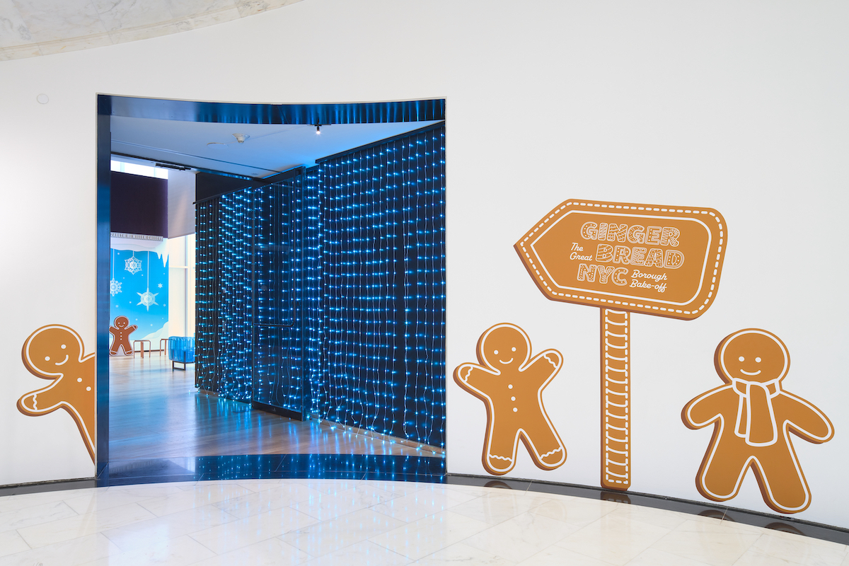 Photograph shows the entrance to the Dinan Miller gallery at the Museum of the City of New York, featuring the installation Gingerbread NYC: The Great Borough Bake-Off. Around the door, which is offset with blue string lights, are three gingerbread people and a sign made to look like a cookie with the exhibition title.