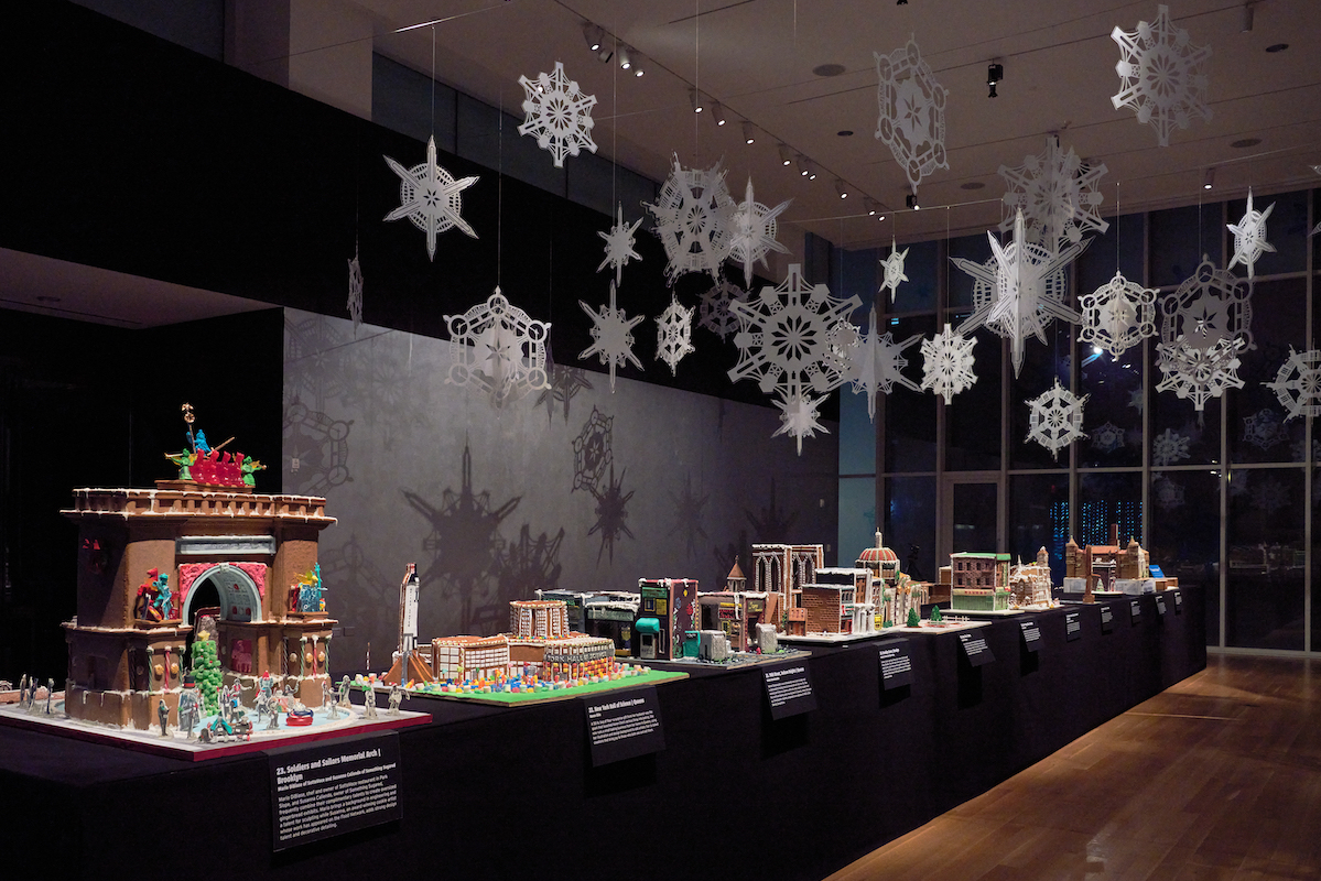 Vue de l'installation montrant certaines expositions de structures emblématiques de New York en pain d'épice exposées dans « Gingerbread NYC : The Great Borough Bake-Off » en 2023. Des flocons de neige sur mesure qui représentent également les structures de New York à la main depuis le plafond et créent des ombres sur le mur.