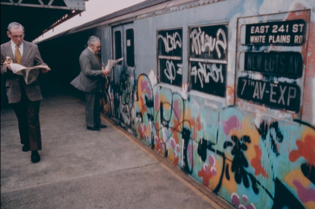Deux hommes plus âgés en costume lisent des journaux sur un quai de métro devant une voiture de métro couverte de graffitis.