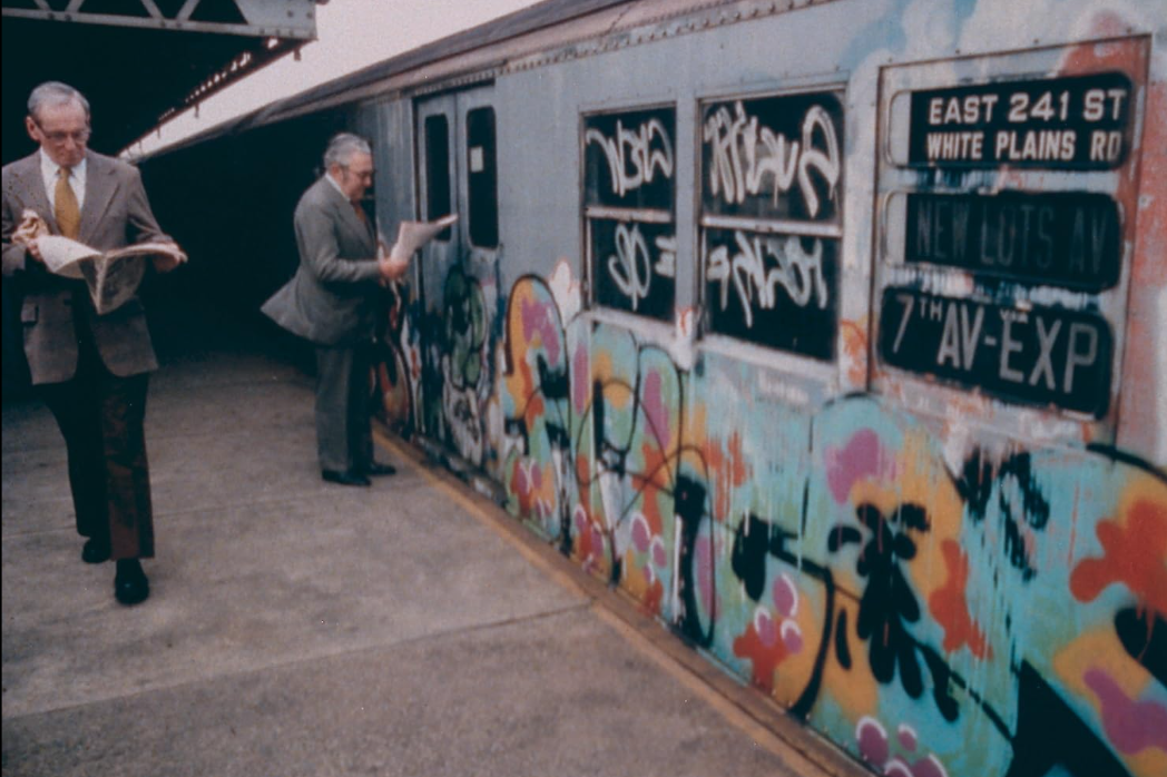 Dois homens mais velhos de terno leem jornais em uma plataforma do metrô em frente a um vagão coberto de graffiti.