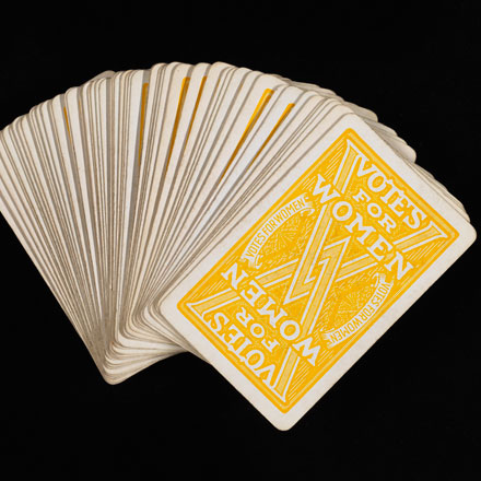 Deck of playing cards. The backs are yellow and say “VOTES FOR WOMEN” with two interlocking “V”s 