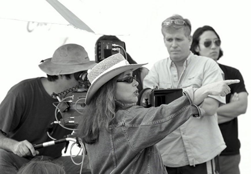 Una imagen en blanco y negro de la directora de cine Susan Seidelman sentada frente a una cámara con un grupo de 3 personas. Ella esta apuntando a la derecha