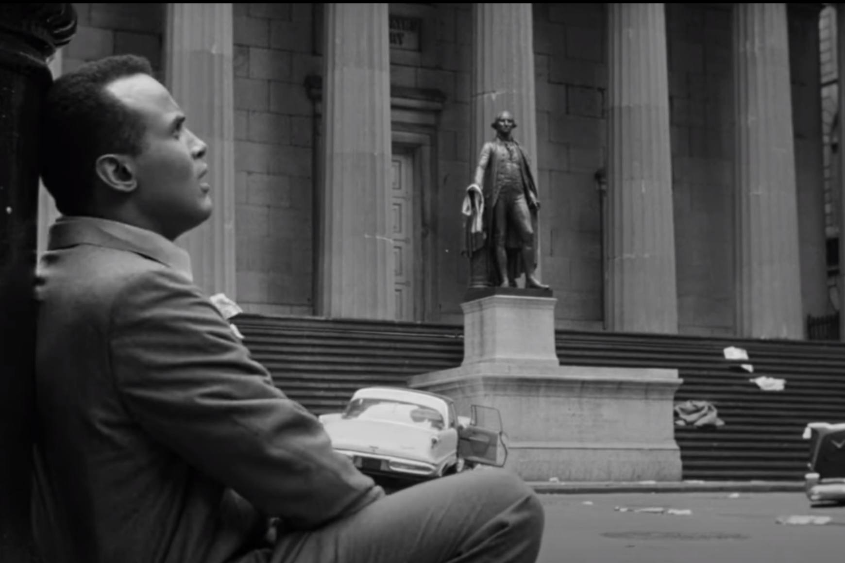 Uma imagem traseira e branca do filme. Harry Belafonte está sentado apoiado em um poste de luz em uma Nova York deserta.