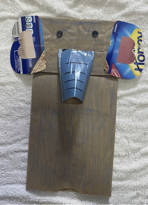 Brown paper lunch bag turned into a hand puppet designed to look like an elephant. It has two black buttons for eyes, two ears made from a blue cardboard food container, and a trunk made from a toilet paper roll covered in blue magazine paper. 