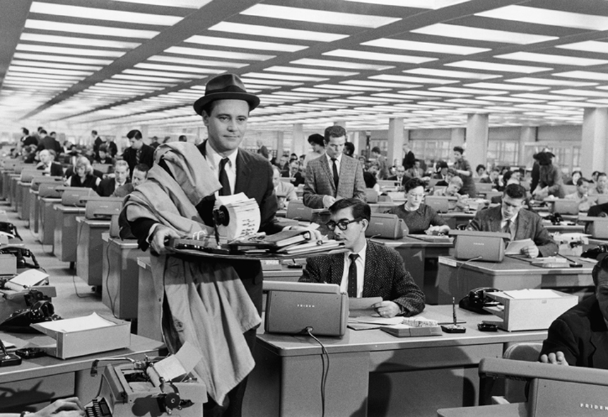 A still from the movie, The Apartment. A man is wearing a suit and holding office supplies, while walking through an office. 