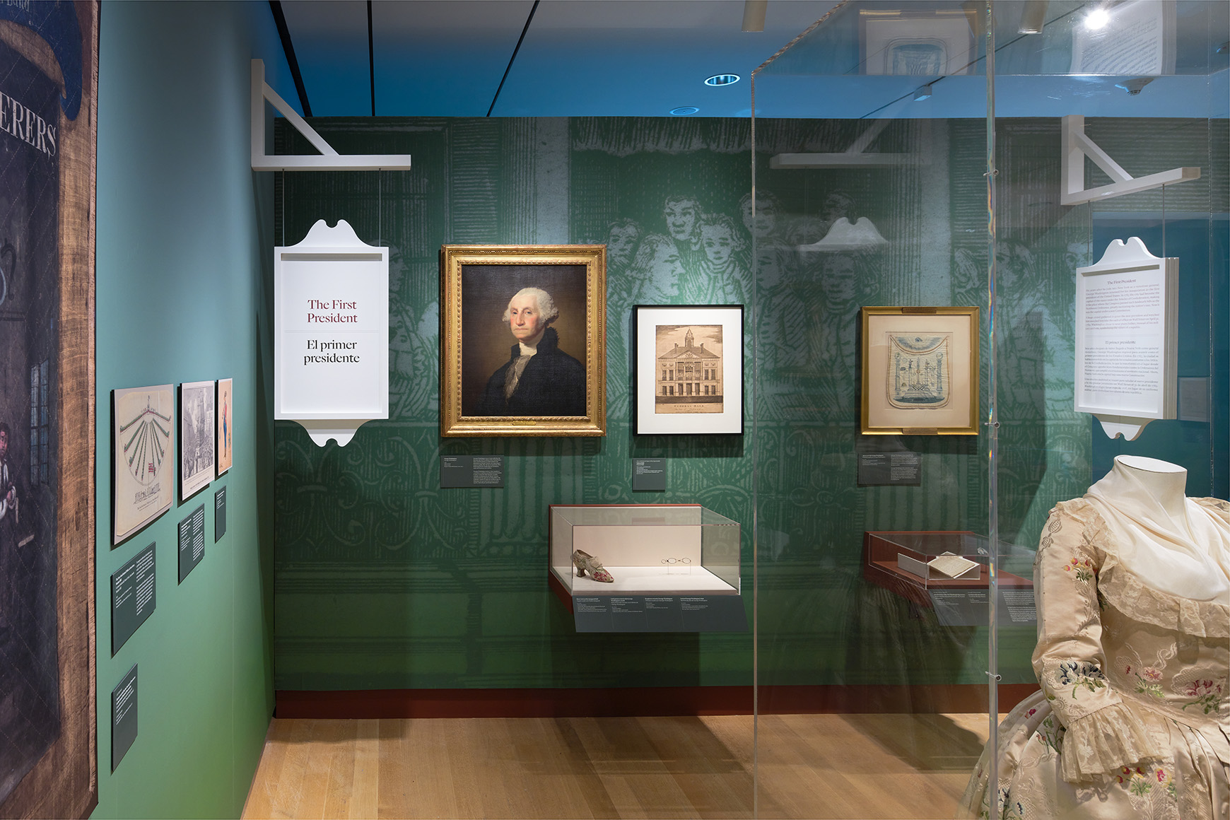 Installation view of the galleries, showing historic clothing, artwork on the walls, and a hanging sign saying, “The First President.”