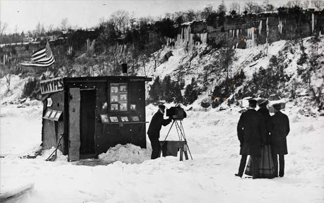 A photographer takes a photograph of a couple in the snow outside of a tintype booth 