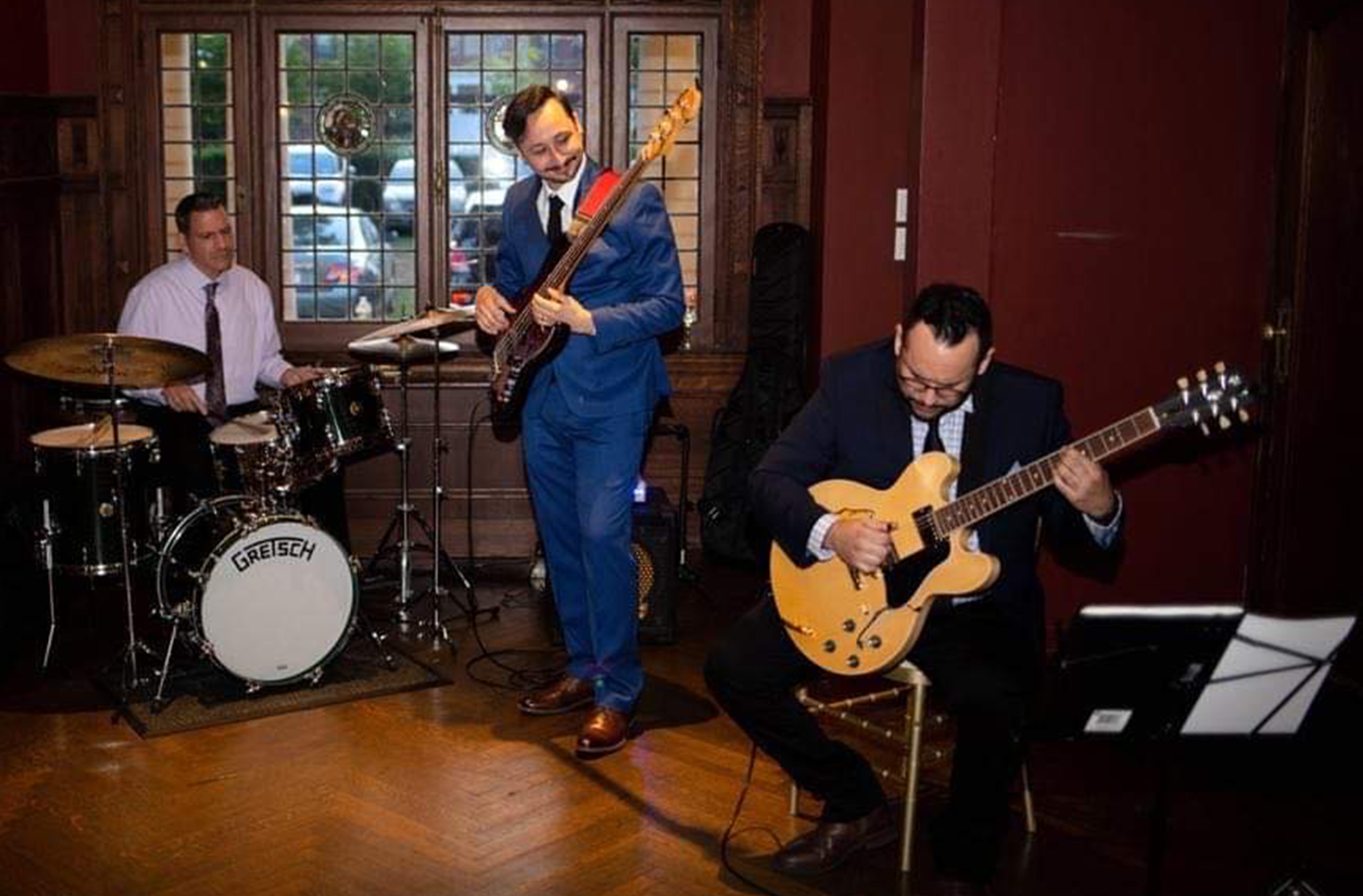 Image of drum kit with drummer, bass guitarist playing a guitar, and electric guitarist sitting and playing electric guitar in a room with a window and cars in the background of the window