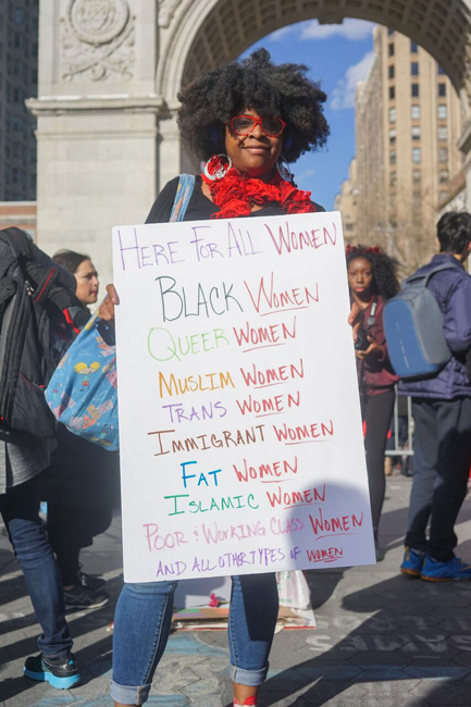 다음과 같은 표지판을 들고있는 여성 : Here For All Women Black Women, Queer Women, Muslim Women, Trans Women, Immigrant Women, Fat Women, Islamic Women, Poor & Working Class Women, and all other types of Women.