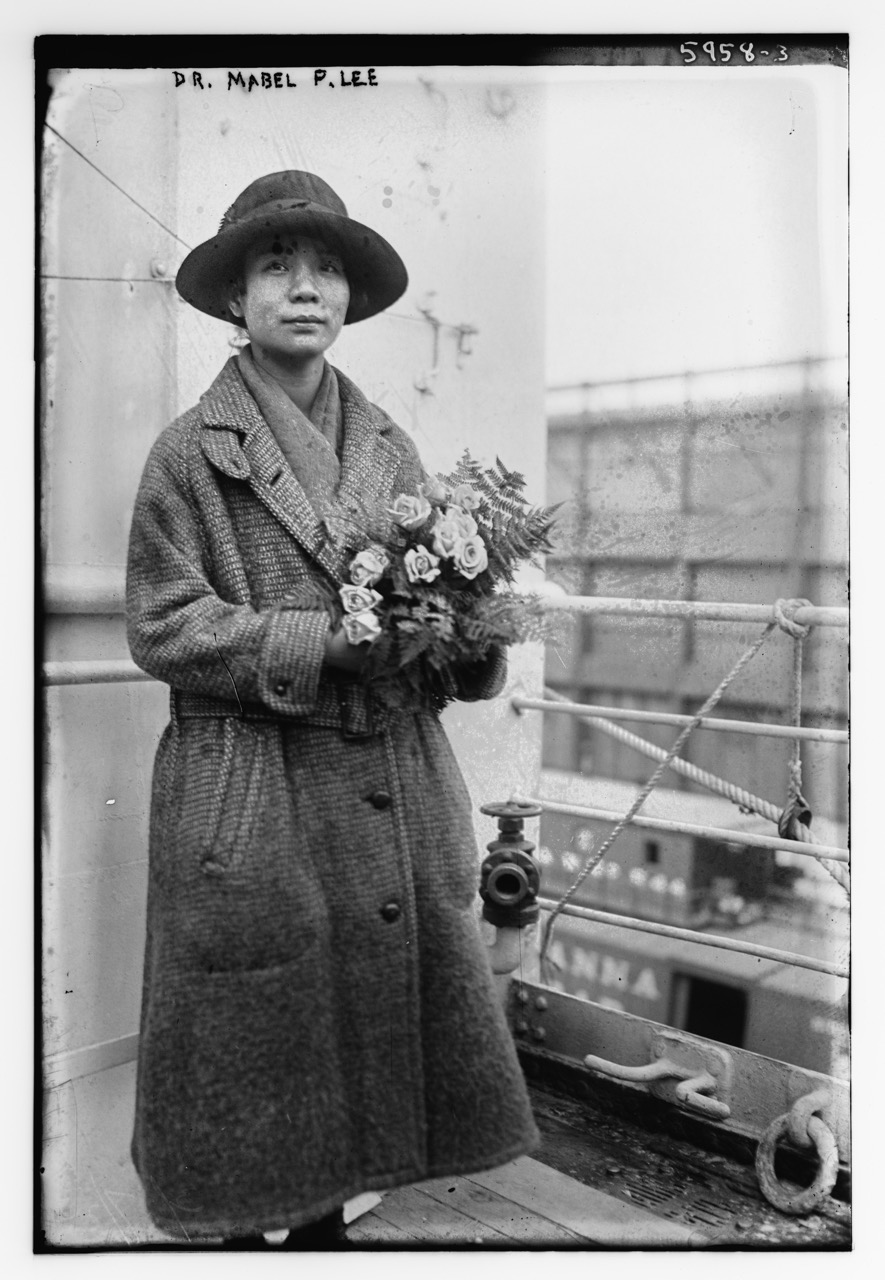 A woman in a hat and large coat holds flowers. 