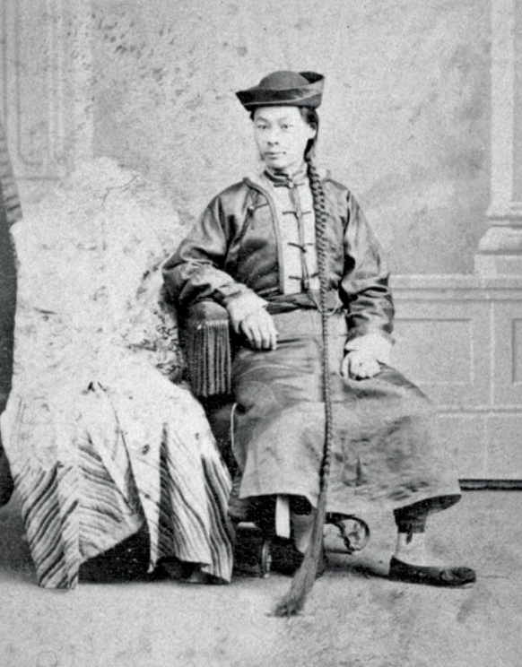 Um homem de chapéu com uma longa trança está sentado em uma cadeira.