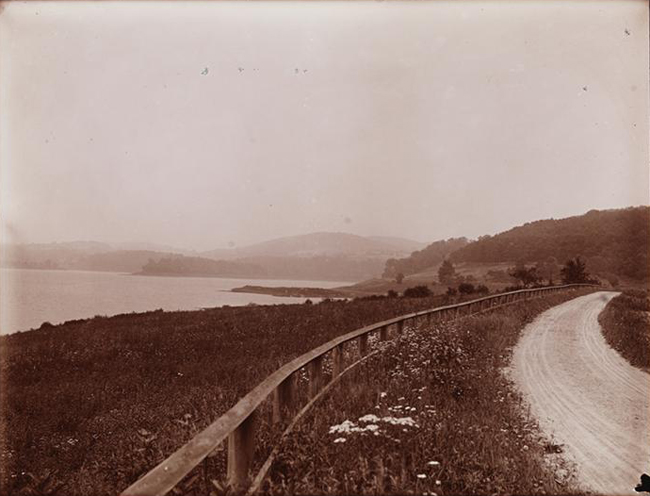 未舗装の道路、クロトン湖に続く丘の中腹、遠くの山々を示す白黒写真。