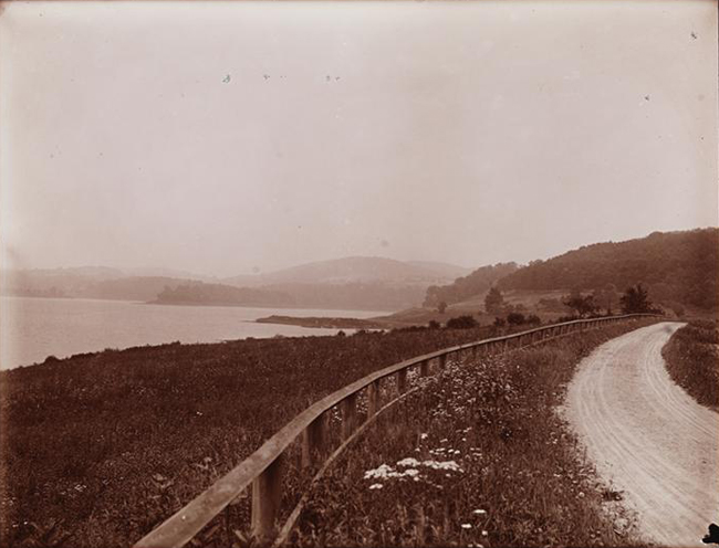 Fotografía en blanco y negro que muestra un camino de tierra, una ladera que conduce al lago Croton y montañas a lo lejos.