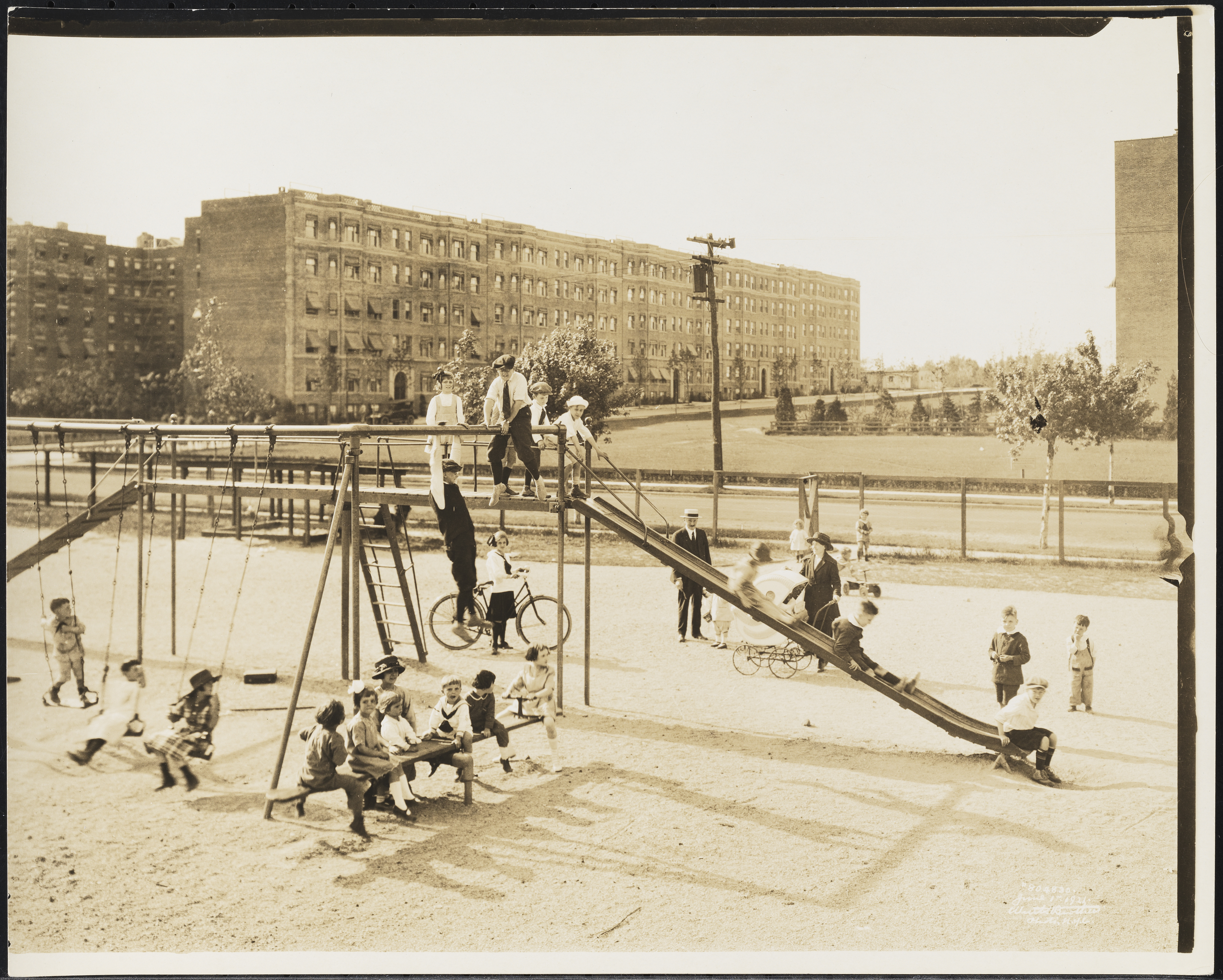 A black-and-white photograph from 1921 showing multiple children playing on a playground with swings, a slide, and a seesaw. Behind the playground is a street and apartment buildings.