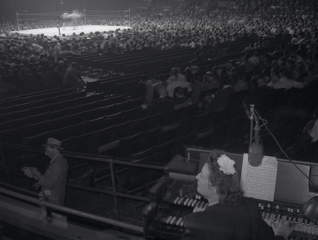 A organista esportiva Gladys Gooding senta-se em um órgão no Madison Square Garden com um ringue de boxe e um público ao fundo.