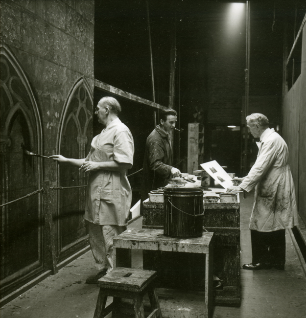Three men in smocks and aprons hold paintbrushes and papers as they paint sets for the Metropolitan Opera with work tables and paint buckets beside them.
