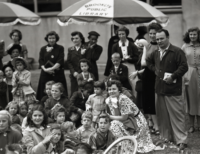 Young boys and girls and some adults sit on the lawn at the Brooklyn Public Library for a summer reading party.