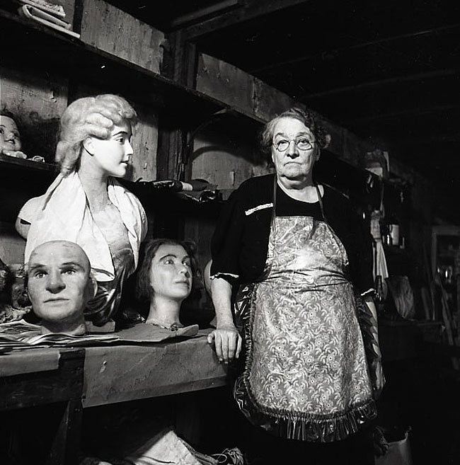 An older woman looks at the camera while surrounded by wax heads she created