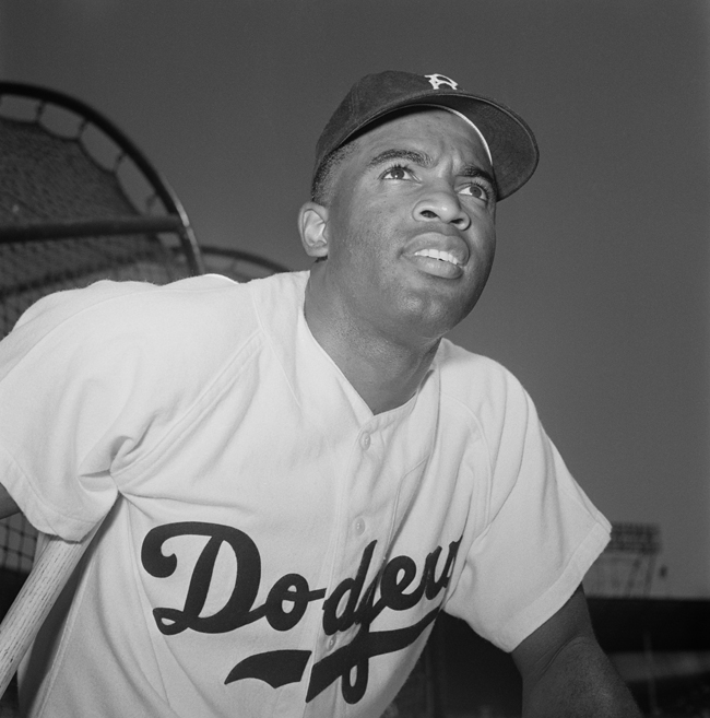 Baseball player Jackie Robinson wears his Brooklyn Dodgers uniform and hat, with a baseball stadium in the background