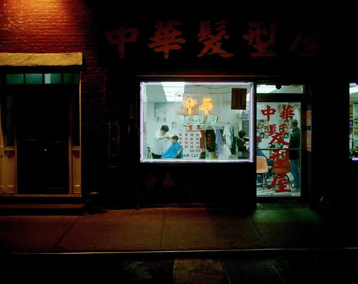 Taken from the street, a boy is seen getting his hair cut in a Chinese barbershop. The signs for the shop are in Chinese.