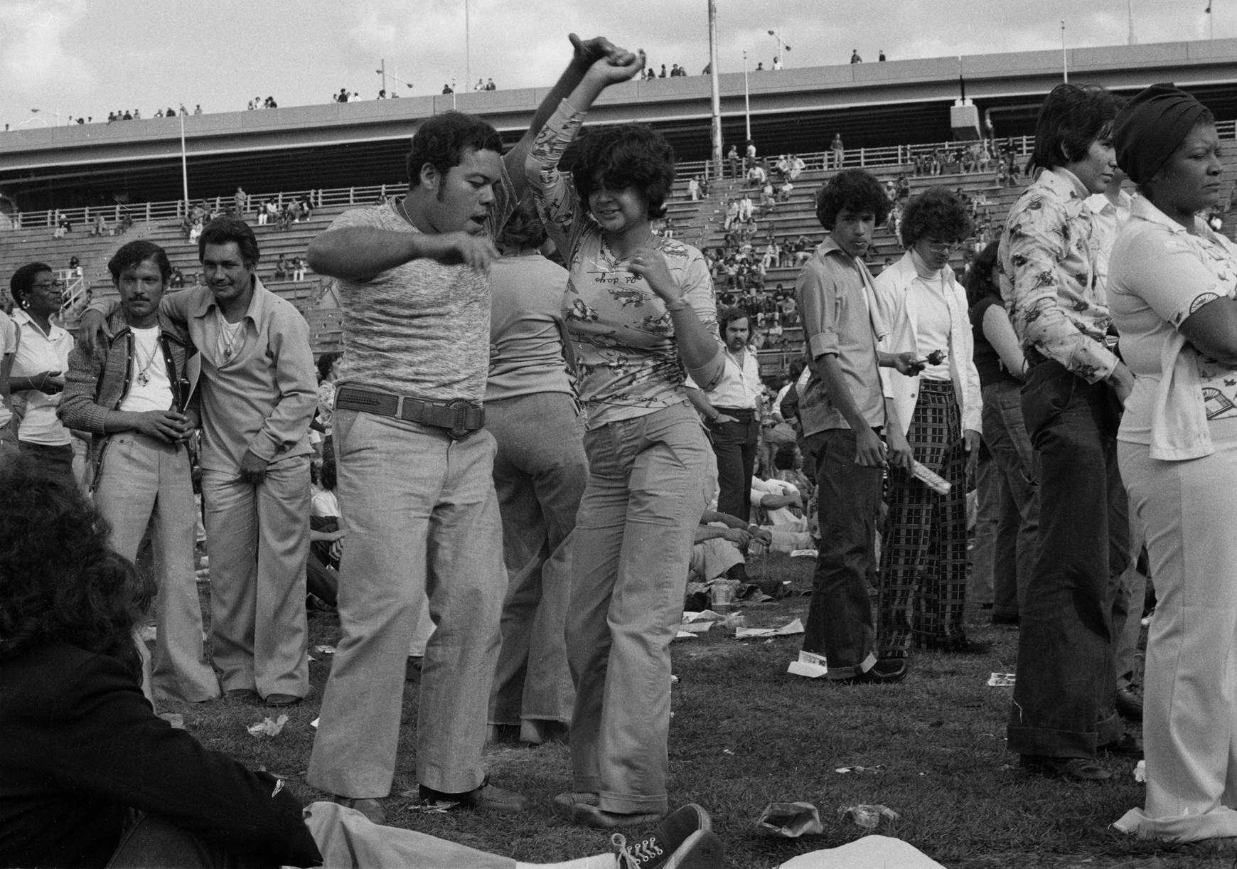 Um casal vestido de maneira casual dança salsa com pessoas ao fundo na Ilha de Randall em 1974.