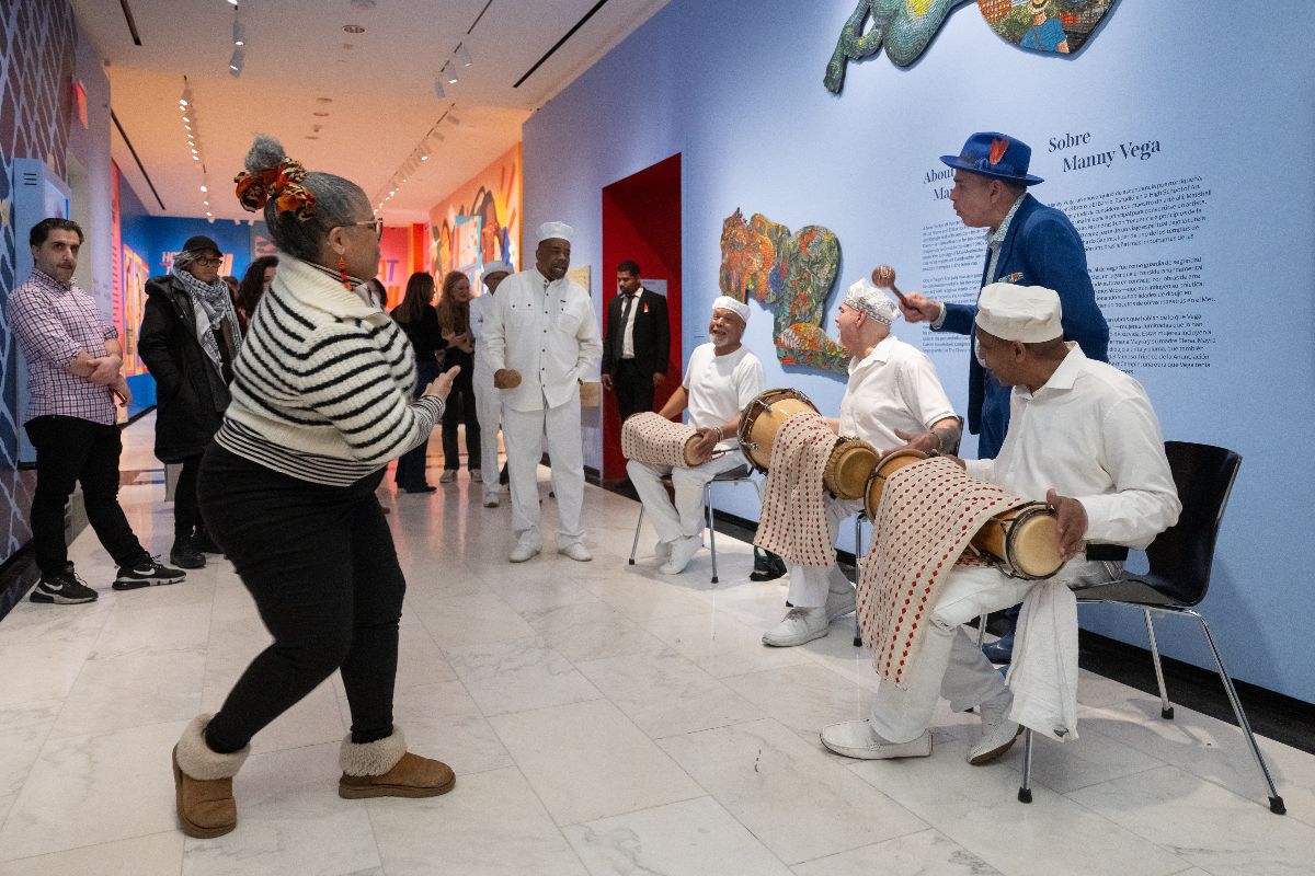 Três músicos vestidos de branco sentados em cadeiras tocando bateria latina. Mulheres dançando na frente de músicos. Artista Manny Vega em terno azul com instrumento shaker.