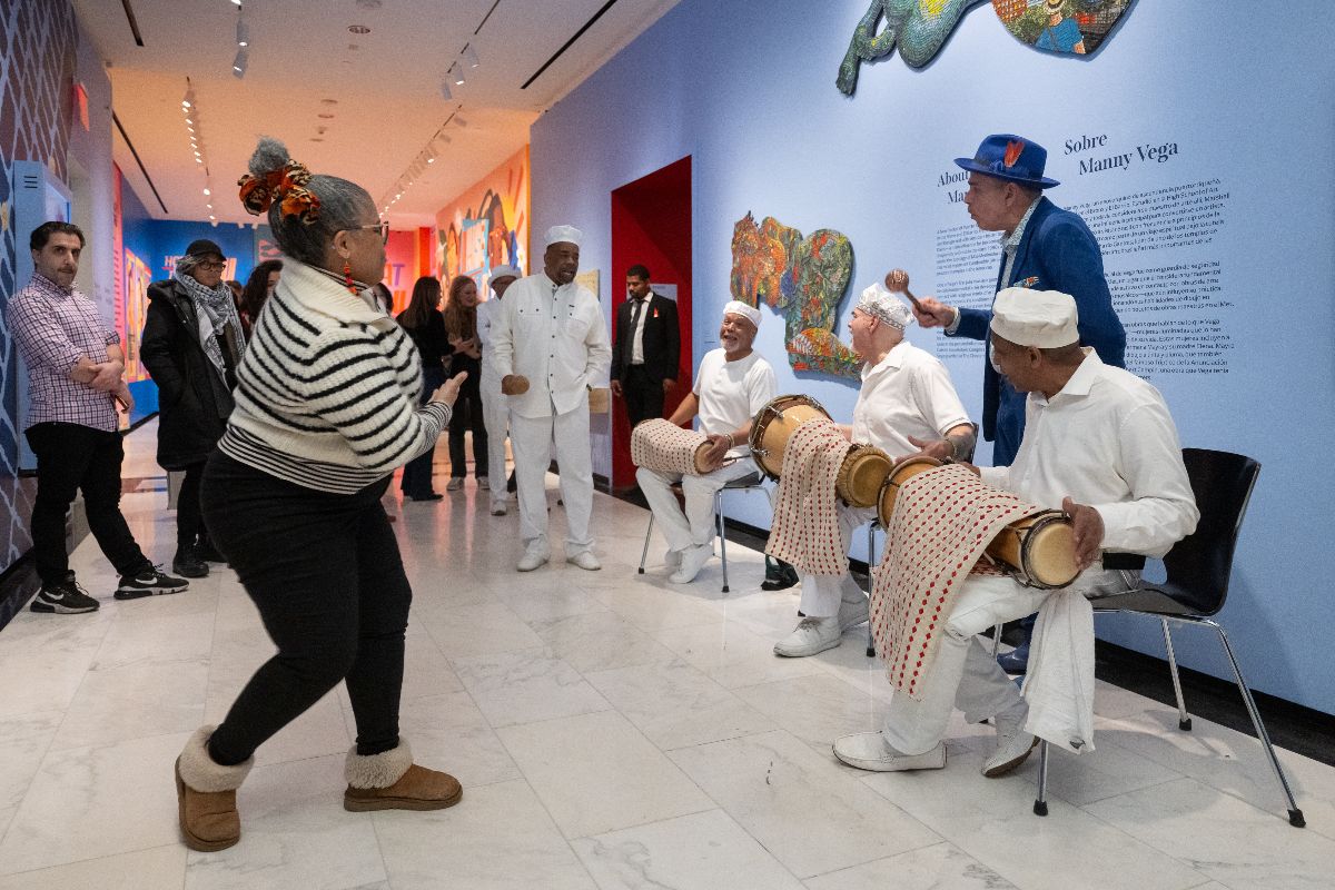 Tres músicos vestidos de blanco sentados en sillas tocando tambores latinos. Mujeres bailando frente a músicos. El artista Manny Vega con traje azul y instrumento agitador.