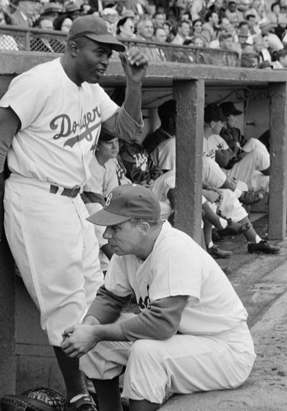 Jackie Robinson se apoya contra el refugio de los Dodgers mientras habla con el capitán Pee Wee Reese