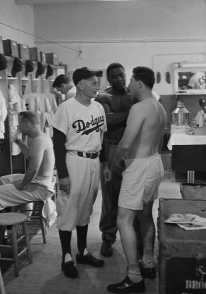 Jackie Robinson은 코치와 팀원과 대화하면서 다저스 클럽 하우스의 라커룸에 서 있습니다.