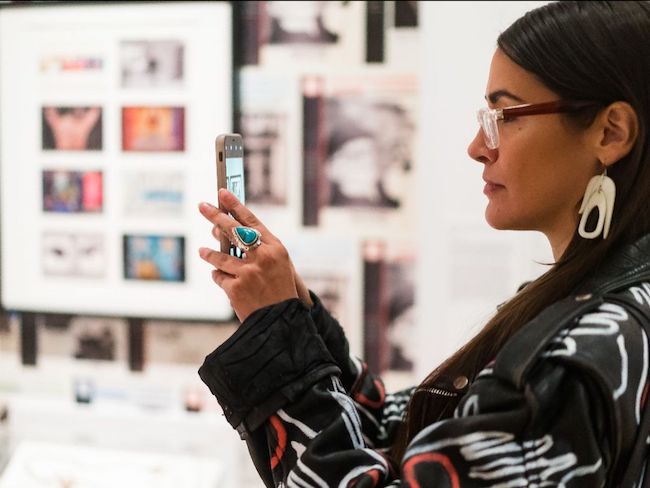 A woman holds her phone to take an image of the exhibition "Urban Indian: Native New York Now."