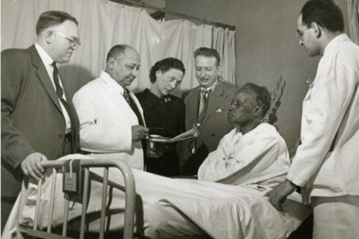 Black and white photograph of Dr. Lyndon M. Hill, Dr. Louis T. Wright, Dr. Myra Logan, Dr. Aaron Prigot, and unidentified hospital employee standing around the bedside of an unidentified African American woman patient.